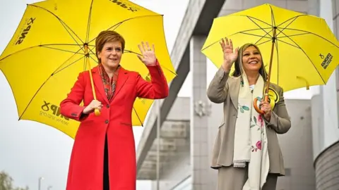 Getty Images Two smiling women with yellow umbrellas wave 