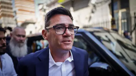 Image shows Andy Burnham wearing black-rimmed glasses and a blue suit, arriving at the Houses of Parliament in Westminster, London, Britain, on 22 June 2026