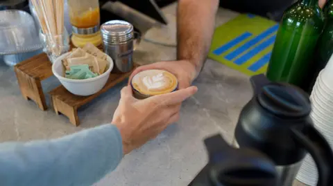 Getty Images an arm in a blue sweater accepts a cappuccino with a leaf decal in the foamed milk