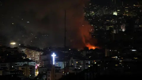 Smoke and orange flames rise, following an Israeli strike on Beirut's southern suburbs. A glowing white cross chan be seen in the foreground 