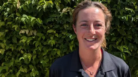 A head and shoulders shot of Jenny Olley, standing in front of a hedge, smiling. She is wearing a navy blue Suffolk Fire and Rescue branded polo shirt.