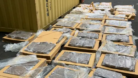 The tobacco is brown in colour and has been packed into rectangle-shaped, vaccum-sealed bags, before being placed in cardboard. The intercepted shipment is laid out on the floor of a warehouse.