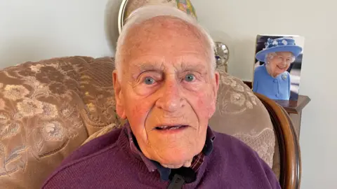 Monty Major sits in his armchair in his front room. He is an elderly man with short white hair. A card with a photograph of Queen Elizabeth, which he received for his 100th birthday, sits on a shelf behind him.