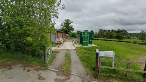 LDRS A brick building next to a small concrete road and green metallic structures behind a silver metal gate in a rural setting