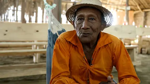 Tito Lopez, community leader of Isbarala, in a bright orange shirt and a straw hat, sit in a swing, look at the camera with a thoughtful expression. Wooden benches and wooden walls and meeting can be seen behind the roof of the house.