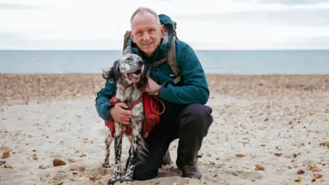 Louis and Nick pictured on a sandy beach, with grey sea visible in the background. Louis has white fur with black markings and black fluffy ears, and wears a red harness and bag. Nick is crouching next to him, wearing hiking books, dark trousers and a dark teal raincoat. 