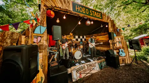 Nathan Roach/Shindig Festival Men stand on a wooden stage and play instruments such as guitars and drums under a sign which says "Hobo stage"
