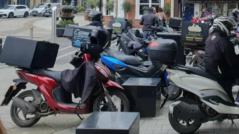 Rebecca Paul A group of mopeds parked on a pavement.