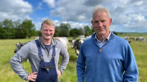 A grey-haired man in a blue jumper and a younger man with a blue boiler suit face the camera with a herd of cows in the background