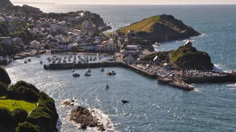 The picture shows a coastal harbour surrounded by steep green hills and a cluster of buildings stretching up the slopes. The water is a deep blue and several small boats are moving in and out of the sheltered harbour area. There are piers curving around the water to protect the boats, and rocky outcrops sit at the harbour entrance where the sea meets the land.