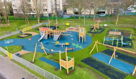 Stevenage Borough Council An aerial photo of an updated play area, showing a pirate ship, climbing apparatus, a zip-line, a swing frame, and blue flooring, around grass areas. A block of flats if behind the play area, where cars are parked, and trees are behind the area too. 