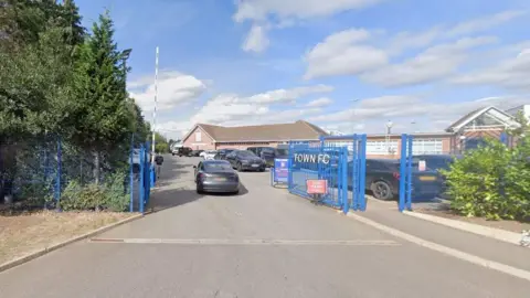 Google Existing entrance to the training ground, showing blue gates, the right hand one of which says "Town FC". There is also a barrier which has lifted to admit a grey car. There are alongside the gates. There is a brick single-storey building in the distance.