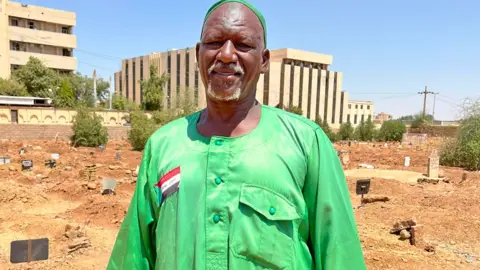 Ken Mungai / BBC Omdurman Gravedigger Abidin Durma berdiri di Pemakaman Sharfi Ahmed - kuburan dapat dilihat di belakangnya. Dia memakai piring hijau cerah, atau khaftan, dengan bendera Sudan dijahit ke atasnya, dan topi kufi dalam warna yang serasi