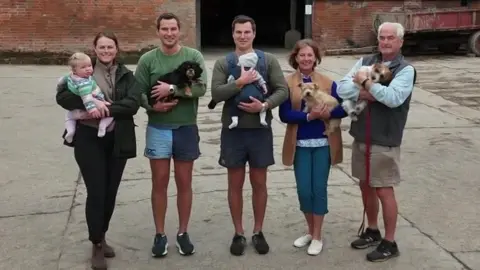 BBC From the left in a line stands Georgie, Tom, Will, Sandra and Peter Carlisle. They are standing in front of their farm buildings on a concrete yard. Georgie is holding her baby daughter and wearing a green coat and black trousers. Tom is holding his black spaniel, wearing shorts and a green jumper. Will is holding his baby in a front carrier and is wearing shorts and a green jumper. Sandra is holding a small dog as is Peter.