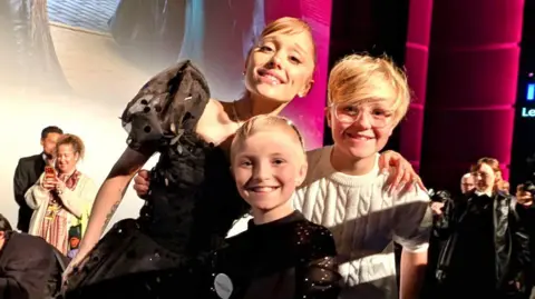 Supplied Two children - including Lola in a black dress - posing with Ariana Grande. All three are smartly dressed and smiling. There is a stage and people behind them.