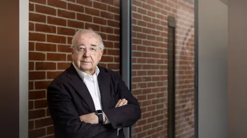 Supplied A man stands smiling at the camera. He has his arms crossed and is wearing a suit jacket and open-neck white shirt. He is standing against a brick wall. 