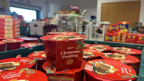 ELLEN KNIGHT/BBC A close-up photograph of a green crate full of pre-packaged Christmas puddings, which have red cardboard packaging with white writing on. Out of focus, in the distance, there are more tables stacked with food like crackers and crisps. 