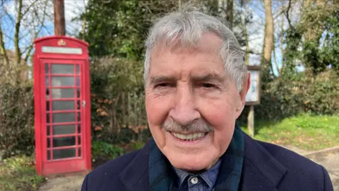 Derek Harris, with grey hair and a grey moustache, wearing a navy coat and blue shirt,  standing beside a red phone box