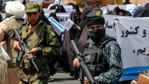 EPA Two armed Taliban soldiers stand guard at a roadside. Both soldiers are wearing camouflage, and one has his face covered. Behind them there is a crowd of people.