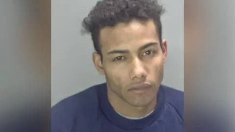 Norfolk Police Police custody photo of Junior Suleimane against a grey background. He is wearing a plain blue crew neck jumper, has short brown curly hair and brown eyes.