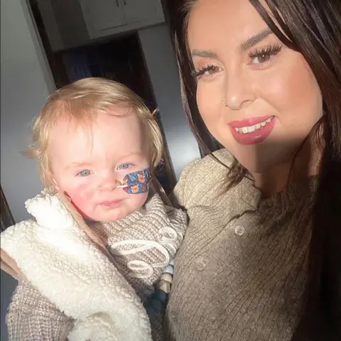 Kristy Evans A little boy and mother smiling. The mother has long brown hair and smiles at the camera. She has a cream jumper on. The boy also has a cream jumper and gilet. He has blonde hair and blue eyes and has a feeding tube through his nose.