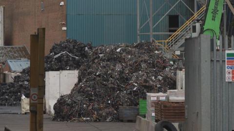 A pile of burnt rubbish at the recycling centre with a steel fence and post in the foreground