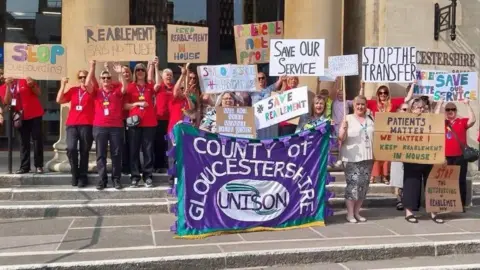 More than a dozen women are standing on the steps of Shire hall. Many have red tops on and black trousers whilst others are dressed in white. They are holding up cardboard placards saying things like "stop outsourcing" and "stop the transfer." The largest banner is a cloth one saying "county of Gloucestershire" on a purple background with the Unison logo below.