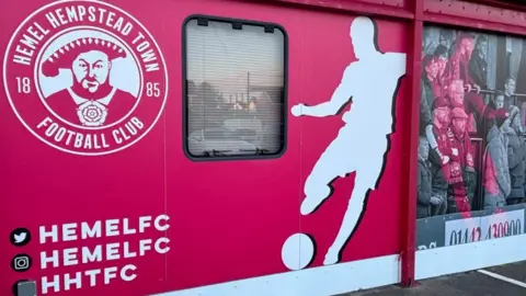 Brian Farmer/BBC A red wall contain a white image of a footballer kicking a ball and a white crest saying Hemel Hempstead Town Football Club. 