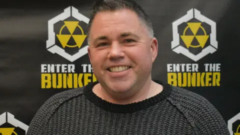 Giancarlo Cininni Jonathan Baha'i, wearing a dark grey sweater, smiles as he stands in front of a wall with radiation symbols and the words 'Enter the Bunker'