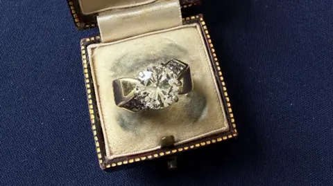 A diamond ring in an open vintage brown ring box on a dark blue background