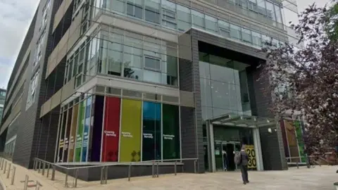 A corner view of a large office block with a paved forecourt. The lower ground floor windows have multi-coloured panels.