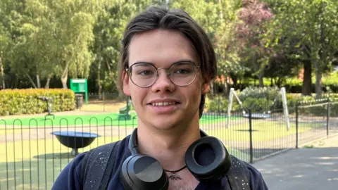 Kevin Shoesmith/BBC A man in his early 20s with sweeping brown hair. He is wearing round-framed galsses and has large headphones around his neck. 