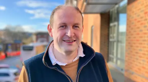 BBC A man with short brown hair is wearing a white shirt, mustard yellow jumper and a navy gilet. He is standing next to a red brick building