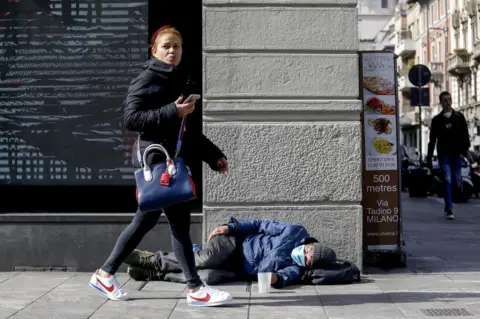 EPA A woman walks by a homeless person who is wearing a face mask