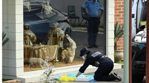 Reuters A police officer is seen on her knees carrying out forensic work outside a window displaying a life-seized nativity scene at the Assyrian Church of the Good Shepherd in Sydney (16 April 2024)