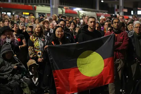 Large group of standing protestors, with several people in the front holding up an Aboriginal flag.