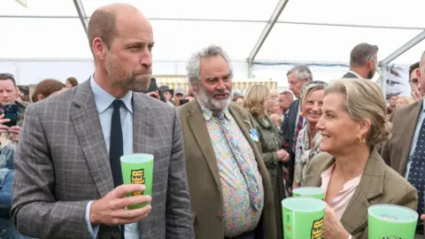 The Prince of Wales at the Royal Cornwall Show with his aunt Sophie, Duchess of Edinburgh. They are standing inside a tent and is surrounded by visitors to the show. They are both trying Rattler cider from a green plastic cup.