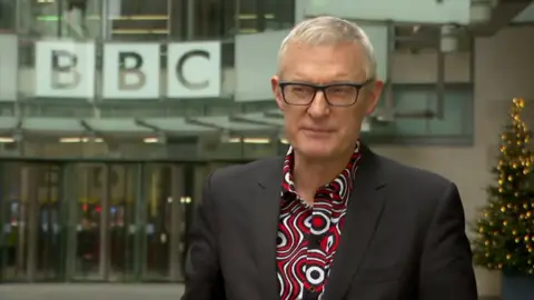 Jeremy Vine stands outside a BBC building, he wears a red and white patterned shirt under a black suit jacket. He has short grey hair and wears black rimmed glasses 