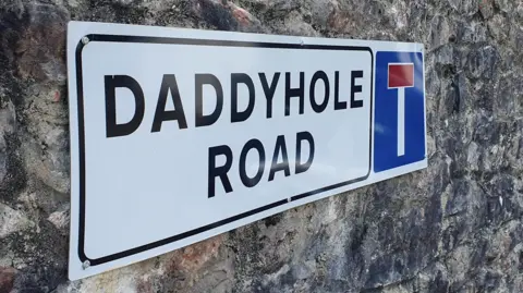 Close up of Daddyhole Road street sign. The white sign is displayed on a stone wall.