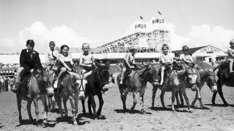 Getty Images Eight donkeys with children on their backs in a black and white photograph dating from 1951. They are on the sand on Coney Beach, Porthcawl, with the fairground including the water chute ride visible in the background.