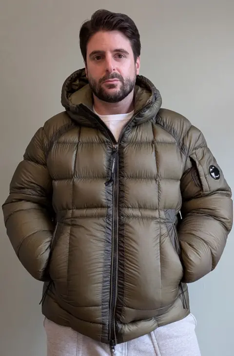 Alex Grossomanides Alex Grossomanides has brown hair, brown eyes and a beard. He is wearing an olive green puffer coat and looking at the camera while standing in front of a white wall indoors.