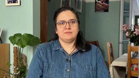 A woman with black hair and glasses wearing denim dungarees