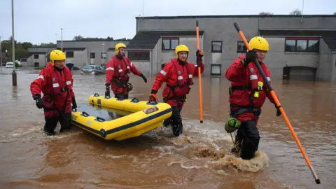 Getty Images A coastguard team walk through a street in Brechin flooded up to knee height. They are in red safety suits and yellow helmets and pulling an inflated craft.