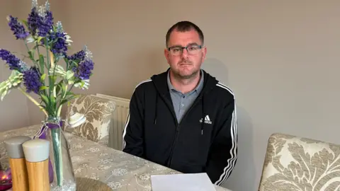 A photograph of a man sat on a table with a floral arrangement to his left and two floral chairs either side of him. The flowers are purple and sit in a vase on the table. Tarran is pictured in a addidas black zip up hoody with white stripes down the arms. He wears a grey top with a collar and has short dark hair and glasses.