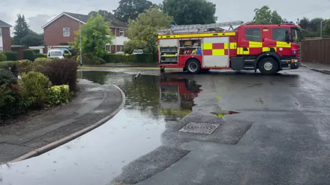 John Devine/BBC A fire engine is parked across a junction in a cul-de-sac. A large amount of water is on the left next to a pavement. A yellow hose is coming from the fire engine and leading down a road. 