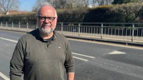 A man wearing a khaki jumper over a black T-shirt looking straight at the camera with a neutral expression. He is standing on a pavement in front of a road with newly installed speed bumps.