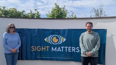 SIGHT MATTERS Cathryn Bradley a woman with short grey hair and a light blue jumper wears sunglasses, and Peter Marshall a man in a long-sleeved grey top with brown hair, they both stand either side of a Sight Matters banner.