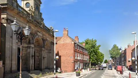 A town centre street, with cars driving on both sides of the road. On the left nearside is a large stone building with arches and Victorian lamppost. Victorian red-brick buildings line both sides of the street further down and there are A-board signs on the pavement on the left.