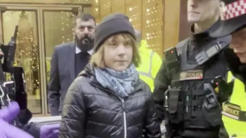 PA Media Greta Thunberg being escorted by police officers outside a building in central London, wearing a black hat, scarf and jacket.