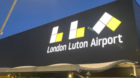 BBC/Danny Fullbrook The front of London Luton Airport. A large black building with a large illuminated LLA and "London Luton Airport" written on it in white font.